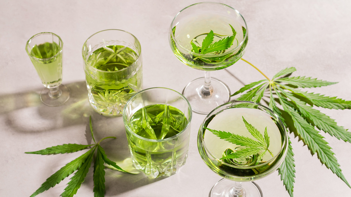 Varios vasos de bebidas con cannabis de color verde pálido decorados con hojas frescas sobre superficie blanca