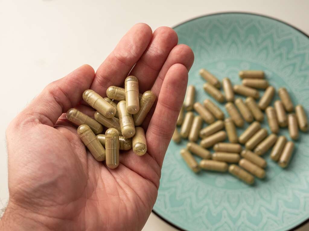 Mano sosteniendo cápsulas de kratom con más cápsulas en un plato verde azulado